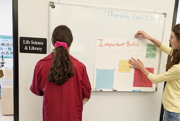 Two students at a whiteboard headed Important British Events and People, beside a door marked Life Science and Library