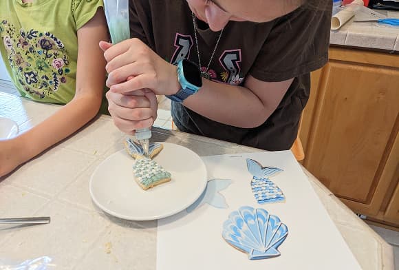 A student piping icing onto a mermaid-tail sugar cookie, with two finished cookies alongside